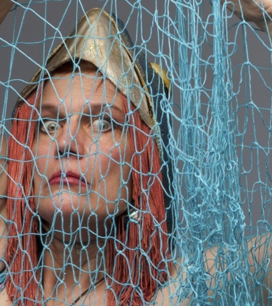 A woman's face, wide eyes, red yarn hair to her shoulders, stares out from behind a blue fishing net.